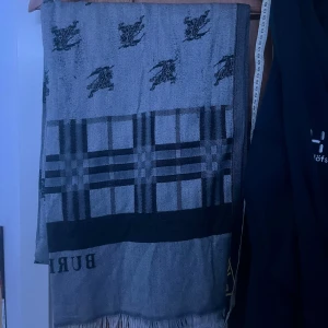 Burberry halsduk med rutor och logga - Supersnygg och lång halsduk från Burberry i grått och svart. Den har klassiskt rutmönster i ändarna och Burberrys ikoniska ryttare tryckt över hela halsduken. Perfekt accessoar för att lyfta din outfit med lite lyxig vibe.Halsduken är 2 Meter lång men går såklart att vika. 2 sidor, en svart, en grå. Har ett litet hål som inte syns!Kom med förslag