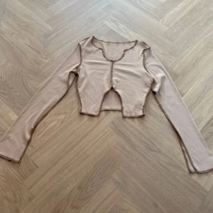 Beige croppad långärmad topp från SHEIN - Säljer en beige croppad topp från SHEIN med lång ärm och ribbad struktur. Toppen har ett randigt mönster och råa sömmar som ger en edgy vibe. Materialet är mjuk polyester och passformen är tight. Perfekt för dig som gillar trendiga detaljer och vill sticka ut.