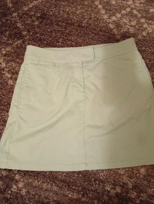 Ljusgrön kortkjol från Puma - Fräsch ljusgrön kortkjol från Puma med klassisk rak passform. Kjolen har praktiska fickor både fram och bak, samt en diskret dragkedja och knappstängning i midjan. Materialet känns lätt och smidigt, perfekt för varma dagar. Puma-loggan är broderad på baksidan. Mint grön