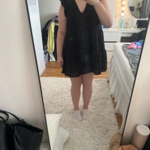 Svart volangklänning - Supersöt svart kortklänning med volangdetaljer vid axlarna och djup v-ringning. Klänningen har en luftig passform och är ärmlös, perfekt för varmare dagar. Materialet känns lätt och skönt!✨💕