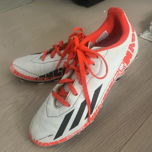 Fotbollsskor adidas  - Säljer ett par Adidas fotbollsskor i vitt med svarta ränder och orange detaljer. Skorna har snörning, platt sula med dobbar och coolt grafiskt mönster längs sidan. Insidan är orange och de har en spetsig tå för bättre bollkontroll. Perfekt för dig som vill sticka ut på planen .