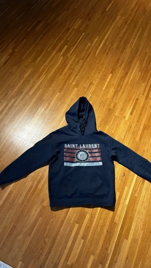Svart Saint Laurent hoodie med tryck - Mörkblå hoodie från Saint Laurent. Väldigt gott skick. Helt ny utan prislapp och kvitto finns tyvärr ej. Därav lite billigare pris än andra! Vid frågor är det bara att höra av dig! Priset är inte skrivet i sten 😃