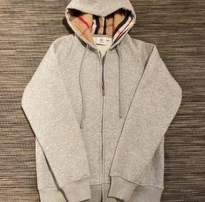 Grå hoodie från Burberry med dragkedja - Snygg grå hoodie från Burberry med klassiskt rutigt foder i huvan. Tröjan har dragkedja framtill, snörning i huvan och ribbade muddar. Perfekt för dig som vill ha en stilren och exklusiv look med en touch av Burberrys ikoniska mönster. Med logo på bröstet 