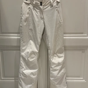 Vita skidbyxor från Helly Hansen - Snygga vita skidbyxor från Helly Hansen med regular passform. Byxorna har dragkedjor vid fickorna, bälteshällor och elastisk midja bak. Materialet är slitstarkt och perfekt för vintern. Helly Hansen-logga på benet för extra stilpoäng.