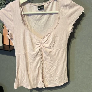 Ljusrosa v-ringad topp från Gina Tricot - Supersöt ljusrosa topp från Gina Tricot med v-ringning och spetskant. Toppen har korta ärmar, snygg rynkning framtill och en liten volangdetalj längs kanterna. Perfekt för dig som gillar romantiska och feminina vibes.