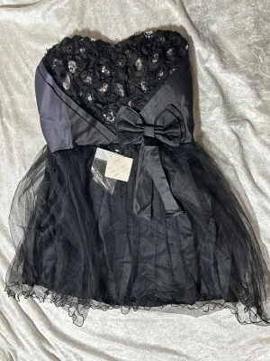 Svart axelbandslös kortklänning med rosett - Svart axelbandslös kortklänning med voluminös tyllkjol och hjärtformad överdel dekorerad med blommiga applikationer och glittriga paljetter. Stor rosett framtill i midjan och smockad rygg för skön passform. Perfekt för dig som vill sticka ut med en glam look. Helt ny 