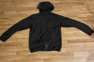 Svart vindjacka från Helly Hansen - Svart vindjacka från Helly Hansen med huva och hel dragkedja framtill. Jackan har långa ärmar, två sidofickor med tryckknappar och meshfoder på insidan. Perfekt för blåsiga dagar och enkel att matcha med din stil.