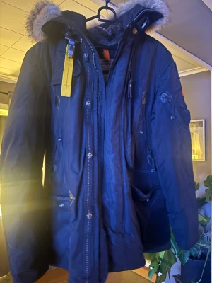Parajumpers parkajacka L - Säljer en svart Parajumpers parkajacka med päls på huvan och ikonisk gul rem. Jackan har flera fickor, patch på ärmen, dragkedja och knappar framtill. Insidan är fodrad med mjuk syntetpäls för extra värme. Perfekt för kalla vinterdagar. Inga skador på varken dragkedjor,knappar eller tyg. Sparsamt använd, byten intressant