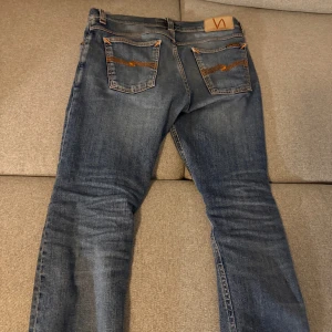 Blå jeans från Nudie tilted tor 30/34 - Säljer ett par snygga blå jeans från Nudie med klassisk tvätt och orange broderi på bakfickorna. Jeansen har slim passform  perfekt för dig som gillar stilrena detaljer och kvalitetsjeans.