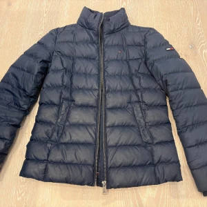 Jacka från Tommy  - Snygg mörkblå pufferjacka från Tommy Jeans med klassisk ståkrage och quiltad design. Jackan har dragkedja framtill, två sidofickor och diskret logga på ärmen. Perfekt för kalla dagar och enkel att matcha med din stil.