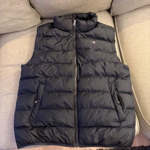 Svart dunväst från GANT - Snygg svart dunväst från GANT med quiltad design och hög krage. Västen har två praktiska fickor med dragkedja och en diskret logga på bröstet. Perfekt att slänga över en hoodie när det är kyligt ute. Stängs med dragkedja framtill.