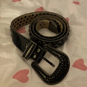 Svart bälte med nitar från B.B. Simon - Säljer ett svart bälte från B.B. Simon med glansig yta och massor av svarta nitar och metallringar. Bältet har en stor, dekorerad spänne och coola detaljer längs hela remmen. Skriv för fler bilder!!
