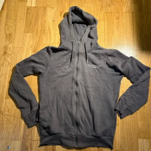 Grå hoodie från McKenzie med dragkedja - Snygg mörkgrå hoodie från McKenzie med huva och snörning. Tröjan har dragkedja framtill, ribbade muddar och ett diskret tryck på bröstet. Perfekt för chill dagar eller när du vill ha en enkel streetstyle-look.