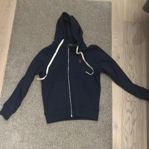 Marinblå hoodie från Polo Ralph Lauren - Snygg marinblå hoodie från Polo Ralph Lauren med vit snörning och dragkedja framtill. Tröjan har huva, två fickor och ett rött broderat Polo-logo på bröstet. Insidan är fodrad med mjuk fleece för extra komfort. Perfekt för chill dagar. Helt ny säljer den pågrund av att den var för liten 