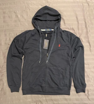Svart hoodie från Polo Ralph Lauren - Svart hoodie från Polo Ralph Lauren med röd broderad logga på bröstet. Tröjan har huva med snörning, dragkedja framtill och två stora fickor. Tillverkad i mjuk bomull, perfekt för chill dagar. Klassisk och stilren design med lång ärm.