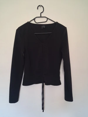 Svart ribbad topp/drawstring crop top Vero Moda - Svart långärmad topp/drawstring crop top från Vero Moda i storlek S. Toppen har ribbad struktur, v-ringning och snörning framtill som kan dras åt för att minska längden på tröjan så att det blir mer som en crop top. Tröjan har några noppror på sig annars är den i mycket bra skick 