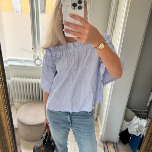 Blårandig offshoulder blus - Säljer en blårandig blus med offshoulder och bred volangkant upptill. Blusen har trekvartsärm och är gjord i ett lätt bomullstyg som känns luftigt. Perfekt för dig som gillar en chill och trendig stil med lite extra detaljer.