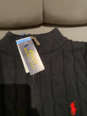 Svart stickad polotröja Polo Ralph Lauren - Svart kabelstickad polotröja från Polo Ralph Lauren med röd broderad logga på bröstet. Tröjan har half zip vid halsen och ribbade muddar. Perfekt för dig som gillar klassisk stil med en modern touch.