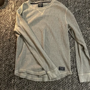 Abercrombie & fitch tröja - Snygg grå långärmad tröja från Abercrombie & Fitch i ribbad bomull. Tröjan har rund hals och ett diskret märke nertill. Perfekt för chill dagar och enkel att matcha med jeans eller joggers.
