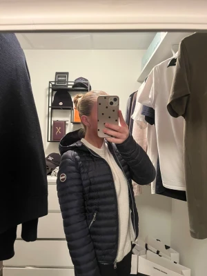 Mörkblå dunjacka från Colmar - Säljer en mörkblå dunjacka från Colmar med huva och dragkedja framtill. Jackan har quiltad design, två fickor med dragkedja och en snygg logga på ärmen. Perfekt för kyliga dagar och riktigt skön att bära. Storlek xs