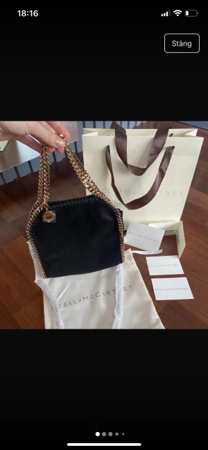 Svart axelväska Stella McCartney - Snygg svart axelväska från Stella McCartney med guldiga kedjor och rund medaljong med logga. Väskan har en mjuk mockaliknande yta och rosa insida med magnetlås. Perfekt storlek för det viktigaste och ger en lyxig vibe till din outfit.