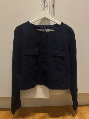 Mörkblå kavajjacka  - Stilren mörkblå kavajjacka med rund halsringning, knäppning framtill och två fickor med lock på bröstet. Jackan har lång ärm och en rak, något croppad passform. Perfekt för en clean och trendig look.