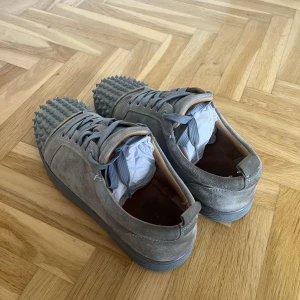 Grå mocka sneakers från Louboutin - Säljer ett par grå sneakers från Christian Louboutin i mjuk mocka med coola nitar på tån. Skorna har rund tå, platt sula och grå snörning. Medföljer extra skosnören och dustbags med Louboutin-logga. Perfekt för dig som gillar edgy designerstil. Bud