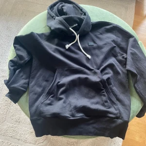 Svart hoodie från Acne Studios - Svart hoodie från Acne Studios med vit snörning i huvan och klassisk känguruficka framtill. Tröjan har lång ärm och en liten svart patch på bröstet. Perfekt för chill dagar och enkel att matcha med.