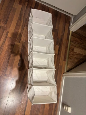 Vit hängande förvaring i tyg - Praktisk hängande förvaring i vit bomull med fem rymliga fack. Perfekt att hänga i garderoben för att organisera kläder, skor eller accessoarer. Enkel att fästa med ögla upptill och smidig att använda i små utrymmen.