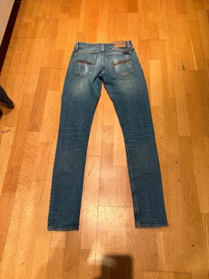 Blå jeans från Nudie Jeans - Säljer ett par klassiska blå jeans från Nudie Jeans med snyggt slitna detaljer och kontrastsömmar. Modellen har fem fickor, tajt passform och raka ben. Jeansen har en ljus tvätt och är tillverkade i mjukt denimtyg med dragkedja och knapp.