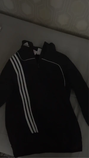 Svart Adidas half zip hoodie - Svart hoodie från Adidas med klassiska vita ränder längs ena axeln och bröstet. Tröjan har en half zip-dragkedja framtill, lång ärm och vit piping som detalj. Perfekt för träning eller chill. Adidas-logga på kragen. Materialet är mjuk bomull.
