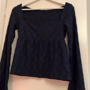 Mörkblå  blus med broderad spets - Säljer en mörkblå blus med långärmade, vida ärmar och fyrkantig ringning. Blusen har ett broderat spetsmönster över hela tyget och en lätt rynkad midja som ger en fin siluett. Perfekt för dig som gillar romantiska detaljer och stilren look.