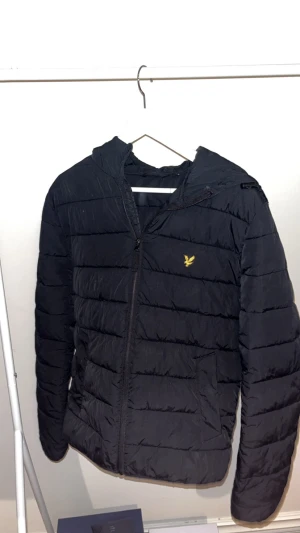 Svart pufferjacka från Lyle & Scott - Svart pufferjacka från Lyle & Scott med gul broderad logga på bröstet. Jackan har huva med snörning, dragkedja framtill och quiltad design. Perfekt för kalla dagar och har en clean, modern look som funkar till streetwear.