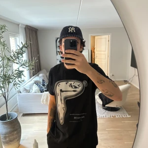 Svart Salvador Dali t-shirt med klocktryck - Svart t-shirt från Salvador Dali x NEUW med stort tryck av en smält klocka inspirerad av 'La Persistencia de la Memoria'. Rund hals, korta ärmar och mjuk bomullskvalitet. Perfekt för dig som gillar konst och unika prints.