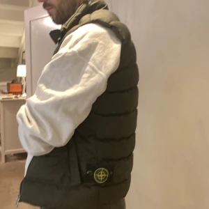 Mörkgrön dunväst från Stone Island - Snygg mörkgrön dunväst från Stone Island med quiltad design och klassisk logotyp på sidan. Västen har dragkedja framtill, ståkrage och två fickor. Perfekt för lager-på-lager och streetwear. Passformen är normal och materialet känns slitstarkt och bekvämt.