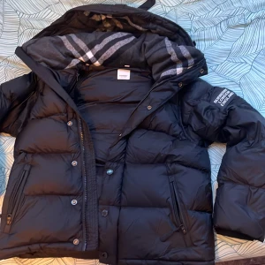 Svart pufferjacka från Burberry - Svart pufferjacka från Burberry med klassiskt rutigt foder i huvan och logotyp på ärmen. Jackan har dragkedja och knappar framtill, samt två sidofickor med dragkedja. Perfekt för kalla dagar med sin tjocka och fluffiga design.