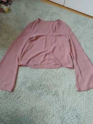 Rosa croppad blus från Ivyrevel - Säljer en ljusrosa croppad blus från Ivyrevel i mjuk polyester. Blusen har v-ringning och långa, lite fladdriga ärmar som ger en chill vibe. Perfekt för dig som gillar stilrena plagg med enkel design och skön passform.