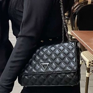 Svart quiltad axelväska från Guess - Snygg svart axelväska från Guess med quiltat mönster och kedjerem. Väskan har en klassisk trekantig Guess-logga framtill och är tillverkad i skinnimitation. Perfekt storlek för att få plats med allt viktigt! 