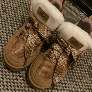 UGG sneakers med päls och snörning - Säljer ett par bruna UGG sneakers med breda mönstrade snören och fluffig vit päls runt öppningen. Skorna har en rund tå, platt sula och detaljer i mocka och syntet. Perfekta för dig som vill ha både stil och komfort under höst och vinter. helt nya bara använt 4 gånger