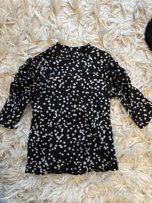 Svart prickig topp  - Svart topp från Lindex i storlek XS med vita prickar över hela tyget. Toppen har trekvartsärm och en högre rundad hals. Materialet är ribbat och känns mjukt mot huden. Perfekt för dig som gillar coola mönster och vill sticka ut lite extra.