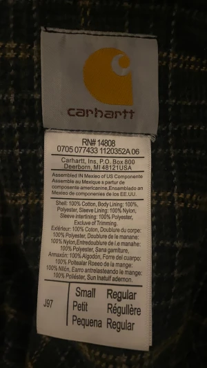 Svart Carhartt bomberjacka - Svart bomberjacka från Carhartt med klassisk krage, dragkedja framtill och brunt logopatch på bröstet. Jackan har två snedställda fickor och en bröstficka med dragkedja. Insidan är fodrad med rutigt tyg för extra komfort. Perfekt för dig som gillar streetwear.