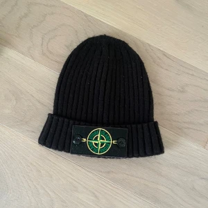 Svart ribbad mössa från Stone Island - Svart ribbstickad mössa från Stone Island med uppvikt kant och klassisk logotyp-patch framtill, fäst med två knappar. Mössan är tillverkad i mjuk ull och har en clean, stilren look som passar perfekt till streetwear.