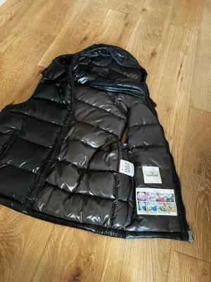 Moncler väst - Snygg svart dunväst från Moncler med glansig finish och avtagbar huva. Västen har två fickor med dragkedja, quiltad design och ikonisk Moncler-logga på bröstet. Insidan har ett coolt serietryck och etiketter. Perfekt för dig som vill ha en trendig look. Storlek M/L