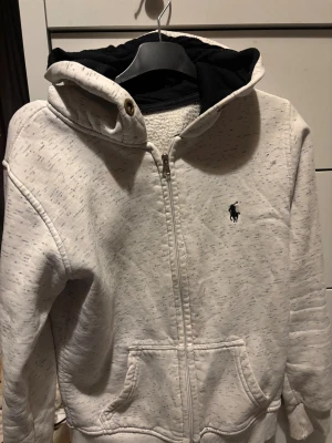 Vit hoodie från Polo Ralph Lauren - Vit hoodie med svarta detaljer i huvan från Polo Ralph Lauren. Tröjan har dragkedja framtill, fickor och broderad logga på bröstet. Materialet är mjukt och känns som bomull, perfekt för kyliga dagar. Långärmad och med ribbade muddar vid ärmslut och nederkant.