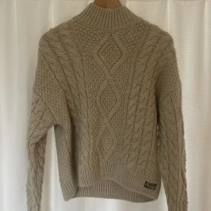 Beige stickad tröja från Superdry - Säljer en snygg beige stickad tröja från Superdry med ett klassiskt flätmönster. Tröjan har en hög krage och långa ärmar, perfekt för kyliga dagar. Den är både stilren och bekväm.