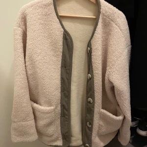 Beige teddyjacka med gröna detaljer - Mysig beige teddyjacka med gröna manchesterdetaljer längs knäppningen och halsen. Jackan har stora fickor framtill och stängs med knappar. Perfekt för dig som gillar chill och comfy stil.