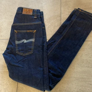 Mörkblå jeans från Nudie Jeans - Snygga mörkblå jeans från Nudie Jeans med klassisk orange söm och vågig broderad detalj på bakfickorna. Modellen har en slim passform och raka ben, tillverkade i slitstarkt jeanstyg. Perfekt för dig som gillar stilrena och tidlösa jeans. 30/32