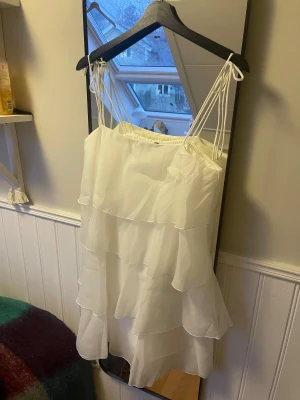 Vit volangklänning  - Supersöt vit kort klänning från Gina Tricot med flera lager volanger och tunna axelband som knyts upptill. Perfekt till sommaren och även studenten! Helt ny, aldrig kommit till användning 
