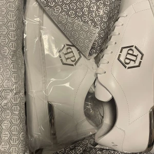 Vita sneakers från Philipp Plein - Snygga vita sneakers från Philipp Plein med en stilren design och metallisk logga på sidan. Skorna har snörning och en diskret dödskalle-detalj på hälen. Perfekta för dig som vill ha en exklusiv look.