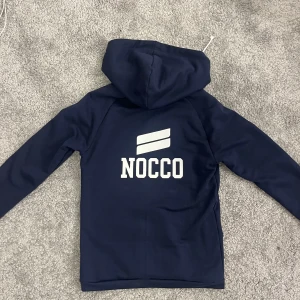 Mörkblå hoodie från NOCCO - Säljer en mörkblå hoodie från NOCCO med vit logga på ryggen. Tröjan har en justerbar huva med snören och en stor ficka framtill. Pris kan diskuteras